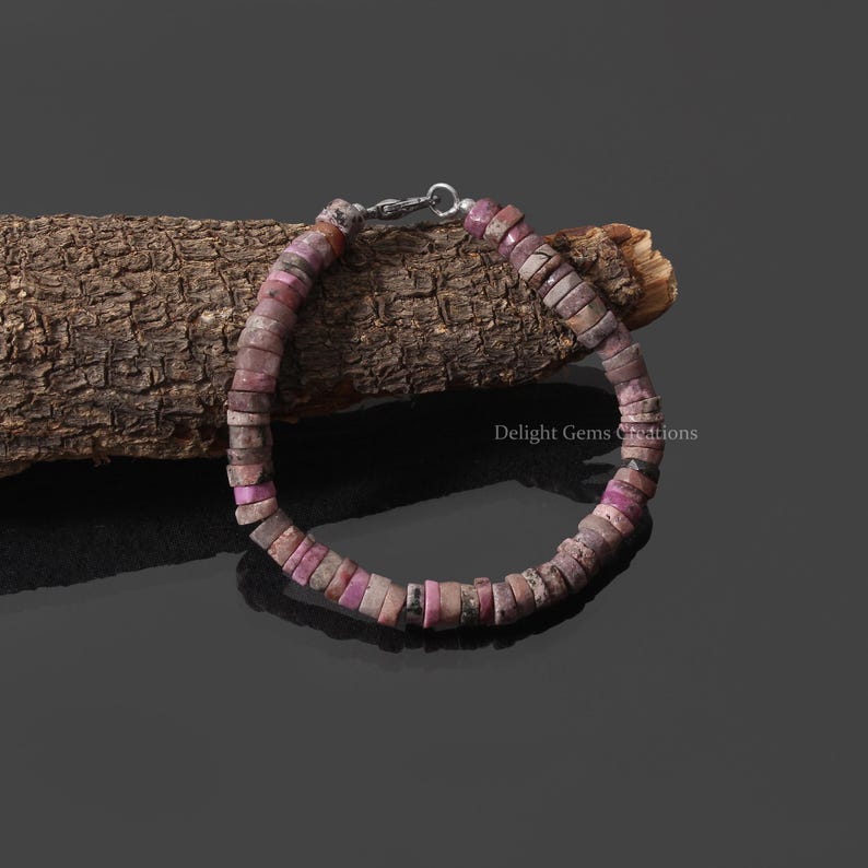 May include: A pink and gray gemstone bracelet with a silver clasp. The bracelet is made of small, round beads and is resting on a piece of wood.