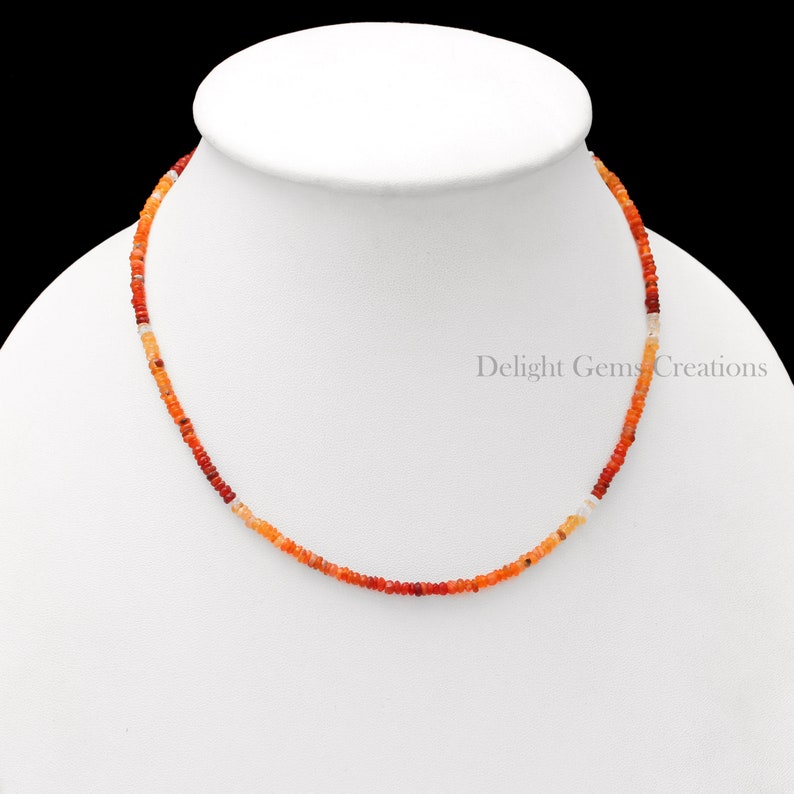 Puede incluir: Un collar delicado hecho de peque&ntilde;as cuentas de &oacute;palo naranja, rojo y blanco graduadas. El collar est&aacute; en un busto de maniqu&iacute; blanco.