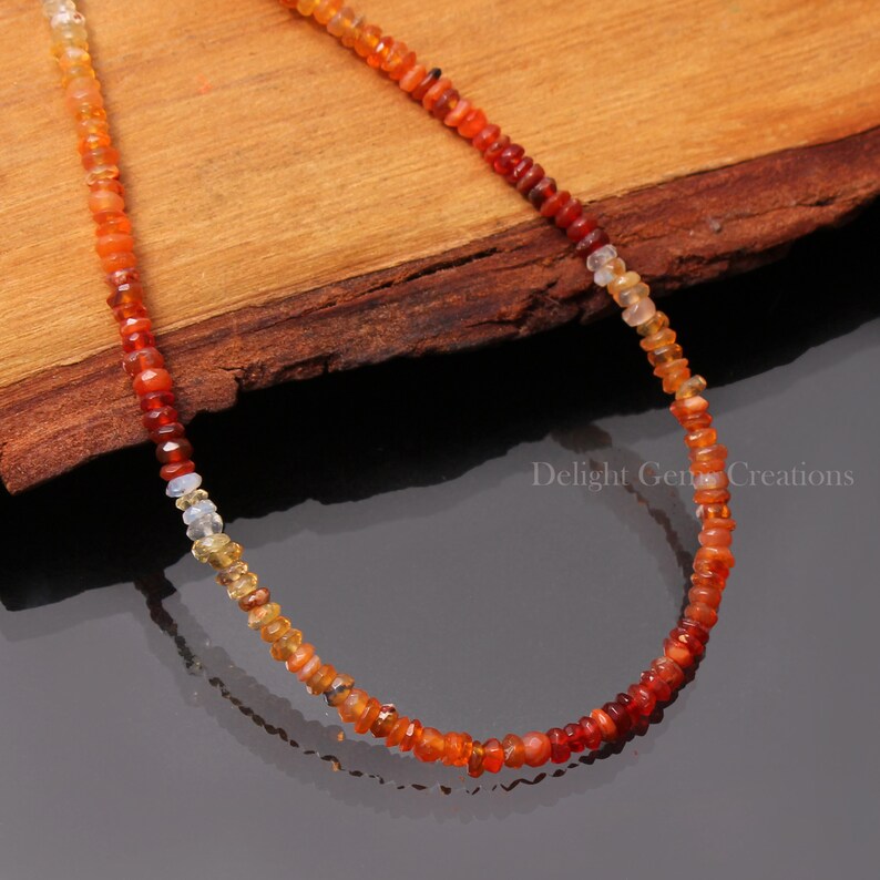 Puede incluir: Un collar hecho de peque&ntilde;as cuentas de &oacute;palo redondas multicolores. Las cuentas est&aacute;n dispuestas en un patr&oacute;n de degradado, comenzando con amarillo claro y pasando a naranja y rojo. El collar se muestra sobre una superficie de madera. Delight Gem Creations est&aacute; impreso en la superficie.