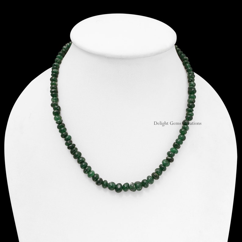 Puede incluir: Un collar de peque&ntilde;as cuentas de piedras preciosas de color verde oscuro. Las cuentas son redondas y est&aacute;n ensartadas. El collar se exhibe sobre un maniqu&iacute; blanco, sobre un fondo negro. Una joya elegante.