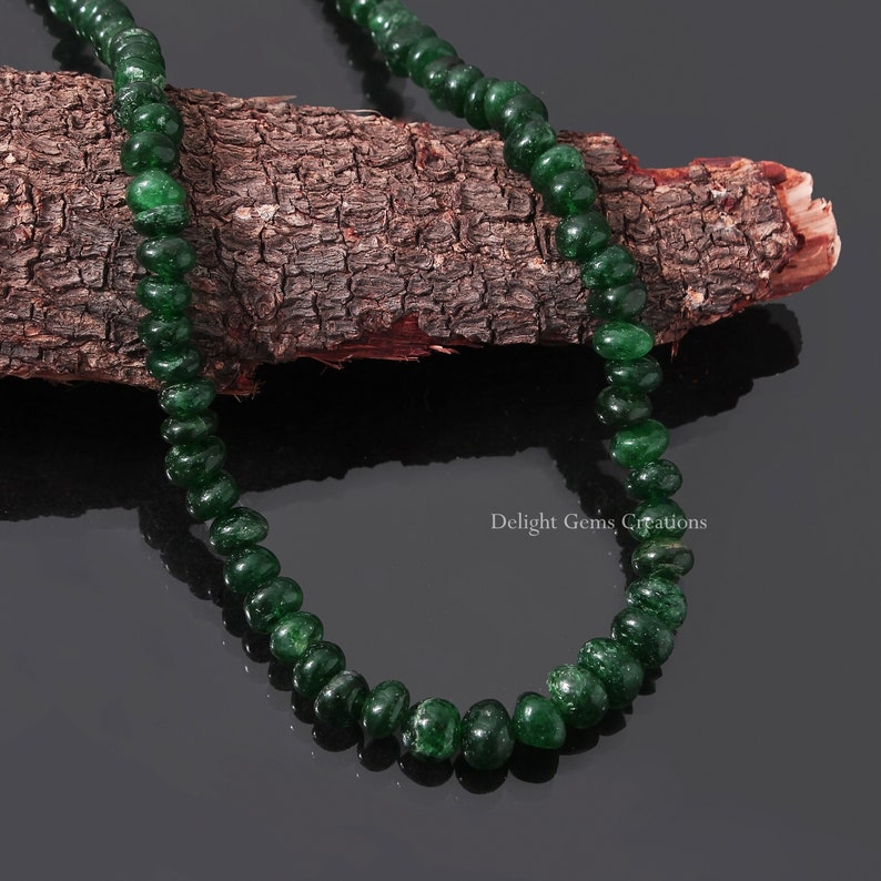 Puede incluir: Un collar de cuentas de piedras preciosas pulidas, de color verde oscuro. Las cuentas est&aacute;n ensartadas, creando una forma suave y redondeada. El collar se exhibe sobre un trozo de madera texturizada sobre un fondo oscuro. El texto "Delight Gems Creations" es visible.