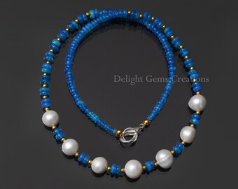 Peut inclure: Un collier en opale bleue et perles. Le collier est composé de petites perles rondes d'opale bleue entrecoupées de perles dorées. De grosses perles blanches sont espacées régulièrement le long du collier. Le fermoir est argenté. Le texte "Delight Gems Creations" est visible.