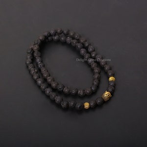 Puede incluir: Un collar de cuentas de piedra de lava negra con detalles dorados. El collar presenta un colgante de cabeza de Buda y cuentas doradas. Las cuentas están ensartadas para formar un collar.