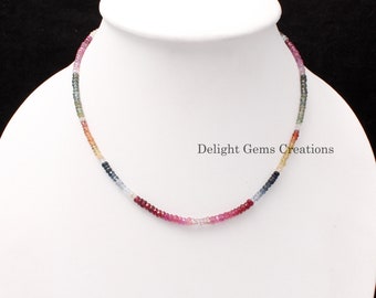 May include: A multi-colored gemstone necklace with small, round beads. The necklace is made of sapphires in shades of blue, green, pink, red, and yellow. The necklace is on a white mannequin bust. The text "Delight Gems Creations" is visible below the necklace.