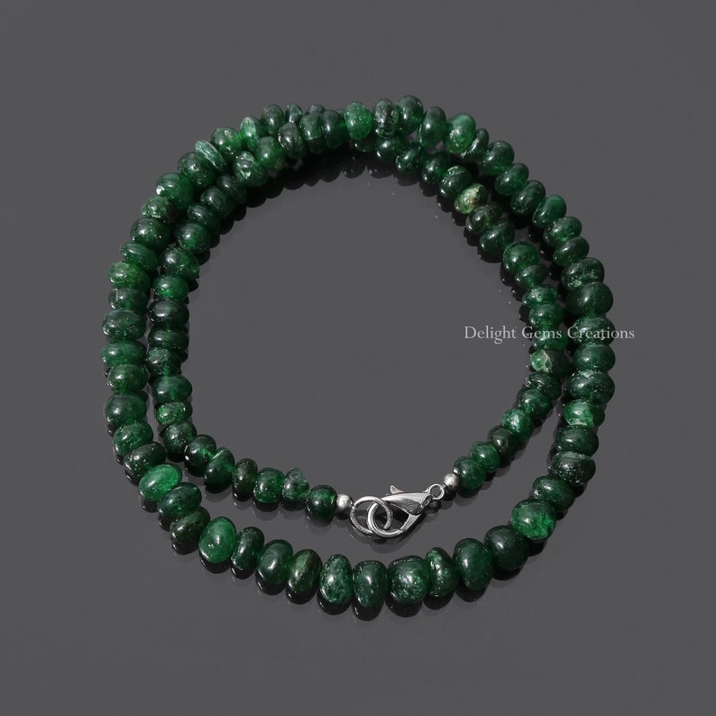 Puede incluir: Un collar compuesto por peque&ntilde;as cuentas redondas de color verde oscuro. Las cuentas est&aacute;n ensartadas, formando un bucle completo con un cierre plateado. El collar se muestra sobre un fondo oscuro.