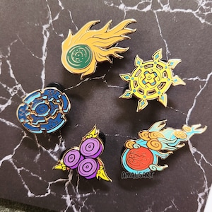 May include: Five enamel pins with gold borders. Designs include a yellow comet with a green centre, a blue and gold circular design, a yellow sun-like shape with blue accents, a cluster of purple circles, and a red orb with blue clouds. The artist's name, "amalsart", is on one pin.