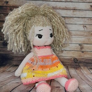 May include: A crocheted doll with blonde curly hair, wearing a pink, orange, and yellow dress. The doll has large brown eyes and a pink face.