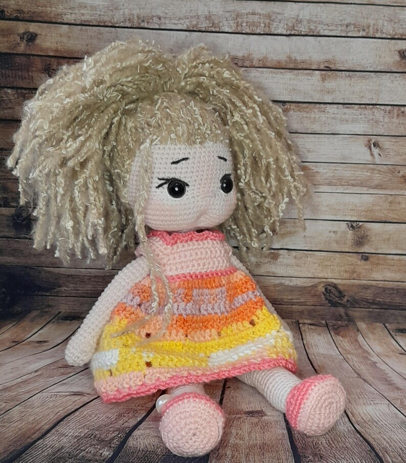 May include: A crocheted doll with blonde curly hair, wearing a pink, orange, and yellow dress. The doll has large brown eyes and a pink face.