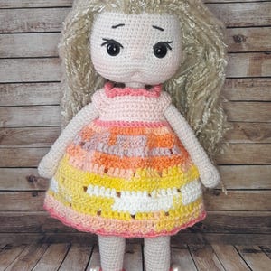 May include: A crocheted doll with long blonde hair, wearing a pink, orange, and yellow dress with white accents. The doll is wearing pink shoes with white pearl accents.