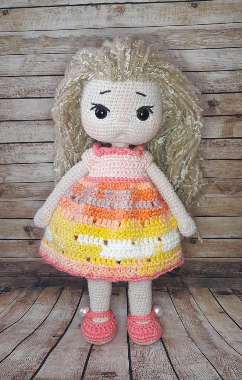 May include: A crocheted doll with long blonde hair, wearing a pink, orange, and yellow dress with white accents. The doll is wearing pink shoes with white pearl accents.