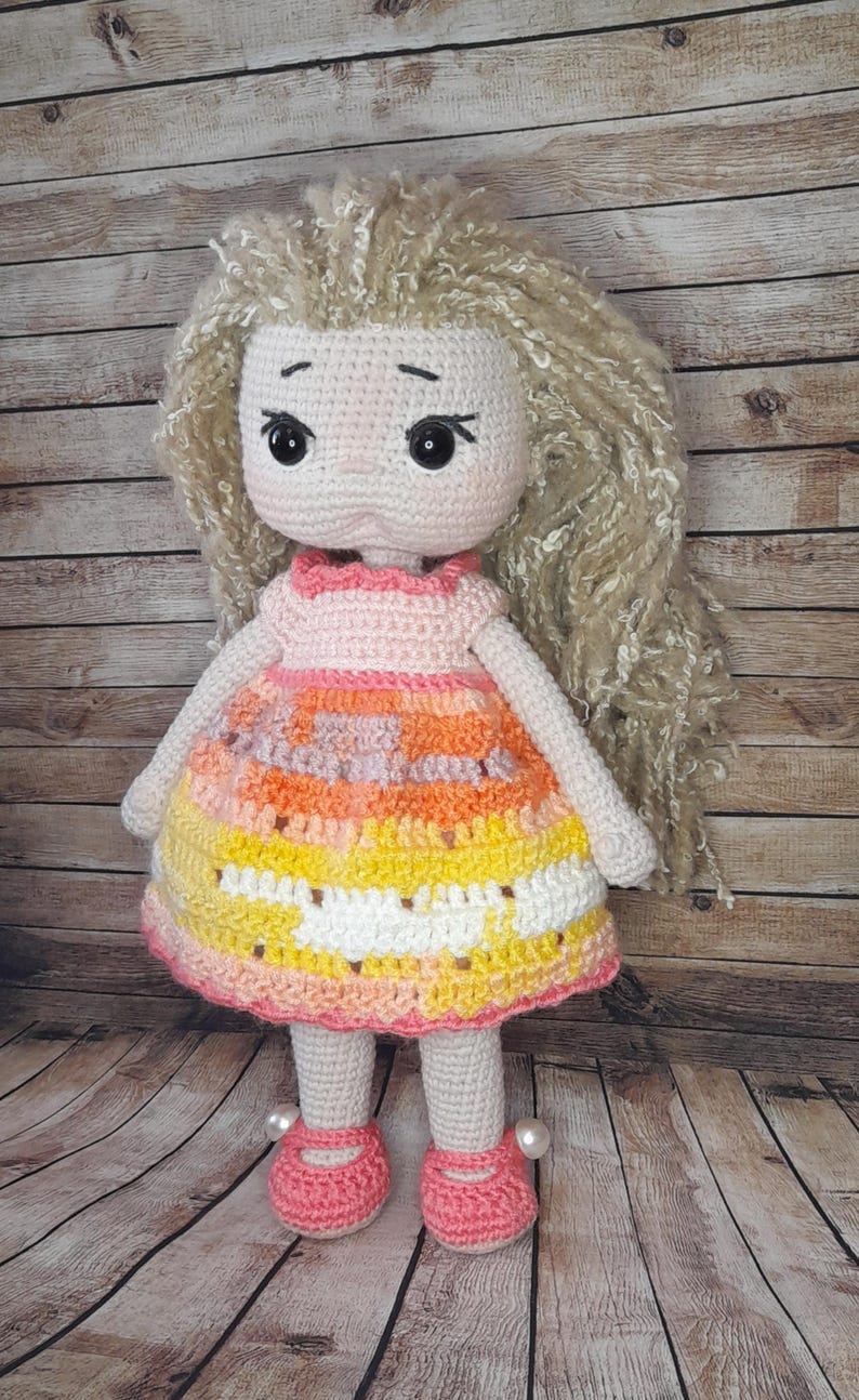 May include: A crocheted doll with long blonde hair, wearing a pink and orange dress with white accents. The doll is wearing pink shoes with pearl accents and is standing on a wooden surface.