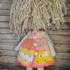 May include: A crocheted doll with long blonde hair, wearing a multi-colored dress with pink, orange, and yellow stripes. The doll is wearing pink shoes with white pearl buttons.
