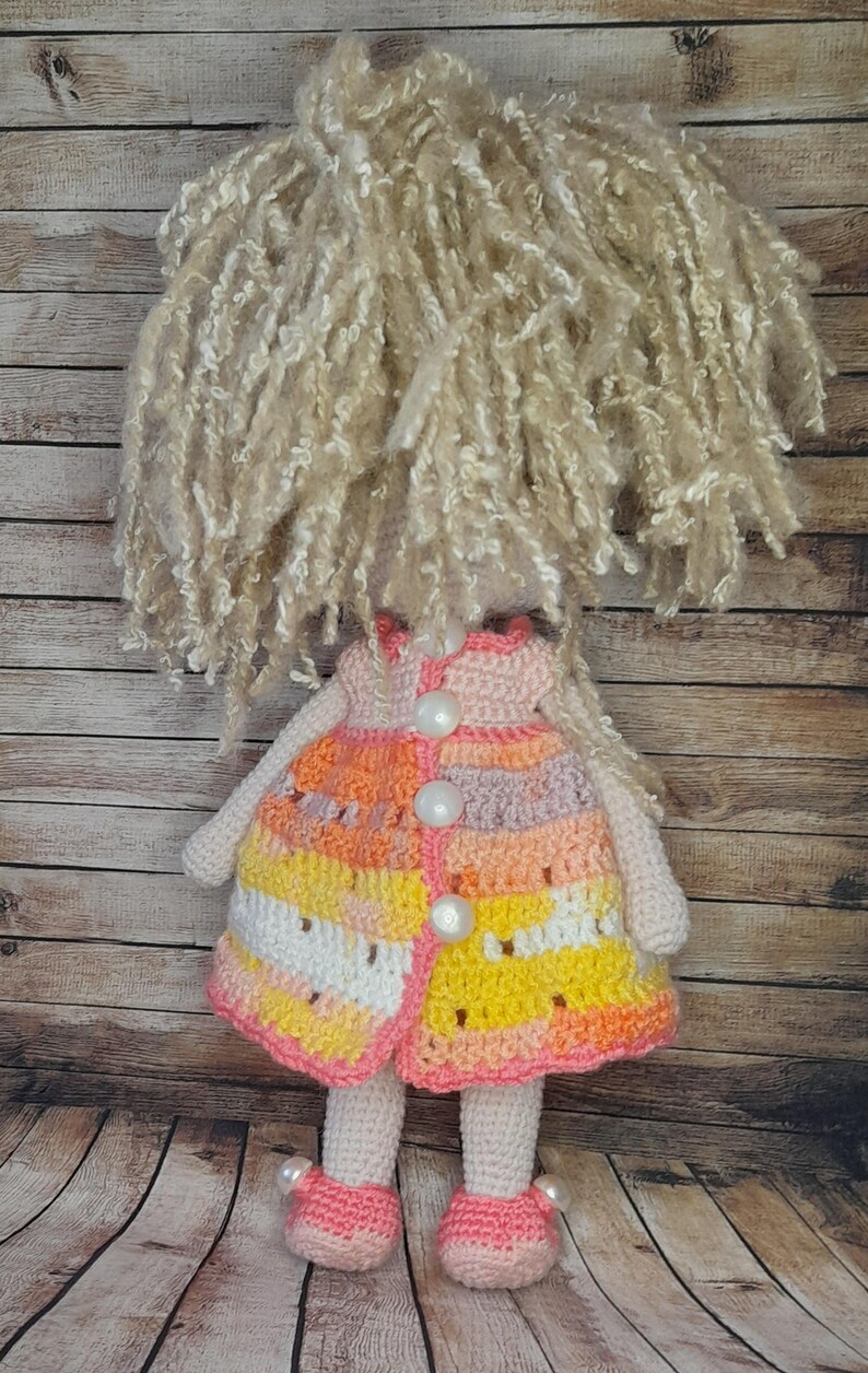 May include: A crocheted doll with long blonde hair, wearing a multi-colored dress with pink, orange, and yellow stripes. The doll is wearing pink shoes with white pearl buttons.