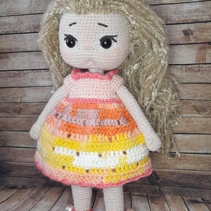 May include: A crocheted doll with long blonde hair, wearing a pink and orange dress with white accents. The doll is wearing pink shoes with pearl accents and is standing on a wooden surface.