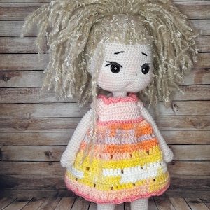 May include: A crocheted doll with blonde yarn hair, wearing a pink, orange, and yellow striped dress and pink shoes.