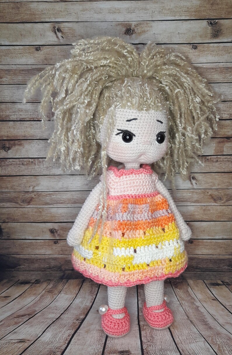 May include: A crocheted doll with blonde yarn hair, wearing a pink, orange, and yellow striped dress and pink shoes.