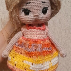 May include: A crocheted doll with long brown hair, wearing a pink and orange dress with white accents. The doll has large black eyes and a pink face. The doll is wearing pink shoes with white accents.