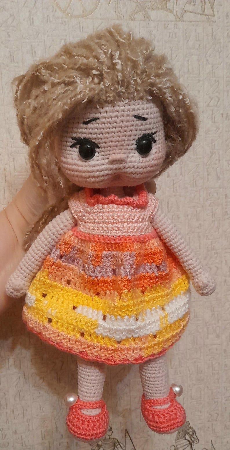 May include: A crocheted doll with long brown hair, wearing a pink and orange dress with white accents. The doll has large black eyes and a pink face. The doll is wearing pink shoes with white accents.