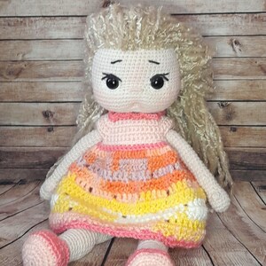 May include: A crocheted doll with long blonde hair, wearing a pink and orange dress. The doll is sitting on a wooden surface.