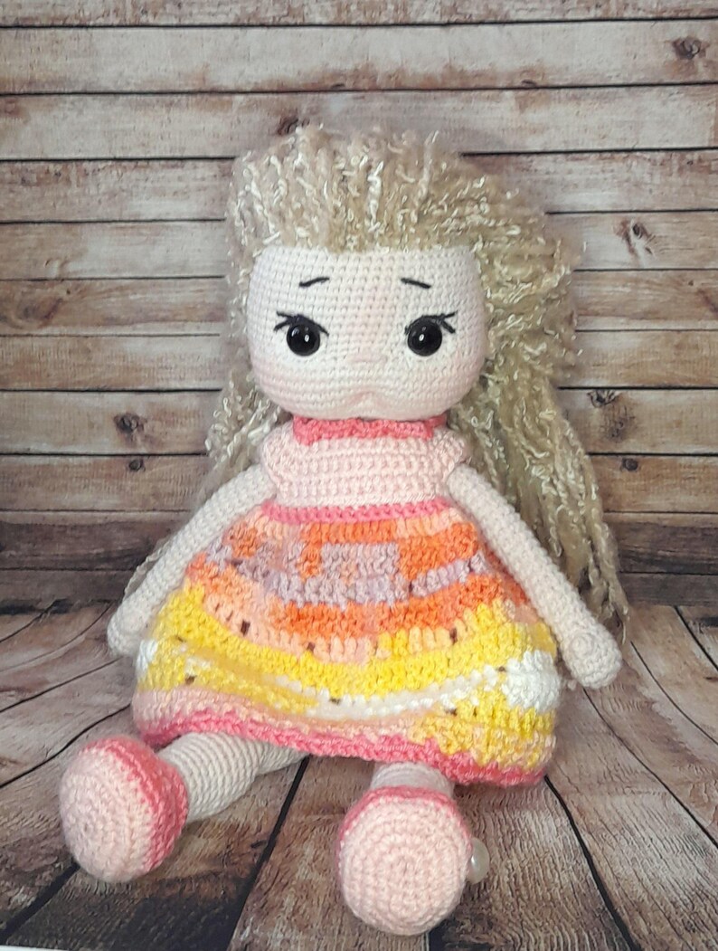 May include: A crocheted doll with long blonde hair, wearing a pink and orange dress. The doll is sitting on a wooden surface.