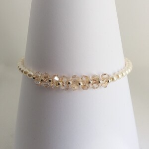 Könnte beinhalten: Ein zartes Armband mit kleinen, klaren Kristallen und weißen Perlen. Das Armband befindet sich auf einem weißen, kegelförmigen Ständer.