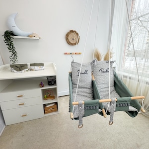 May include: A baby swing with two seats, suspended from the ceiling. The seats are green with gray patterned cushions, and the text "Double Trouble" is printed on the cushions. The swing has wooden bars and gray pom-pom trim.