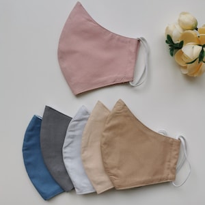 Op de afbeelding: Zes gezichtsmaskers in verschillende effen kleuren, waaronder poederroze, blauw, grijs, lichtgrijs, beige en bruin. Elk masker heeft witte elastische oorlussen. De maskers zijn gerangschikt op een wit oppervlak met een klein boeket crèmekleurige bloemen.