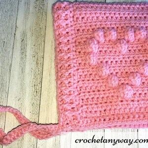 May include: Pink crocheted baby hat with a textured design and a long tie. The hat has a square shape with a raised pattern.