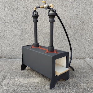 May include: Black metal forge with two burner heads and a brick firebox. The forge is connected to a gas line with a valve and hose.