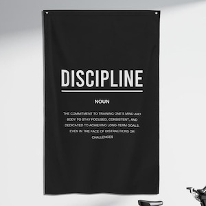 Pu&ograve; includere: Un poster motivazionale in bianco e nero con la parola "DISCIPLINE" in grandi lettere bianche. Il poster definisce la disciplina come "l'impegno di allenare la mente e il corpo per rimanere concentrati, coerenti e dedicati al raggiungimento di obiettivi a lungo termine, anche di fronte a distrazioni o sfide."