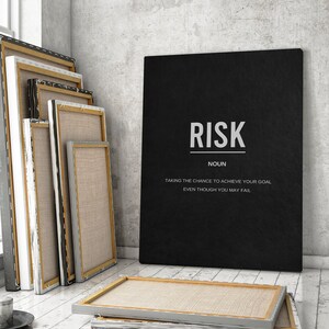 May include: A black canvas print with the word "RISK" in white, accompanied by a definition. Several blank canvas frames lean against a wall and window. The room has a minimalist, modern aesthetic with neutral tones.