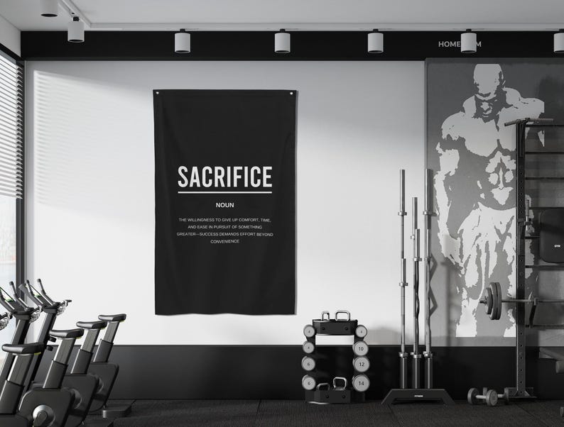 Pu&ograve; includere: Un poster motivazionale in bianco e nero con la parola "Sacrifice" in grassetto in lettere bianche. Il poster definisce il sacrificio come "la volont&agrave; di rinunciare al comfort, al tempo e alla facilit&agrave; nella ricerca di qualcosa di pi&ugrave; grande". Il poster &egrave; appeso a una parete bianca in una palestra domestica.