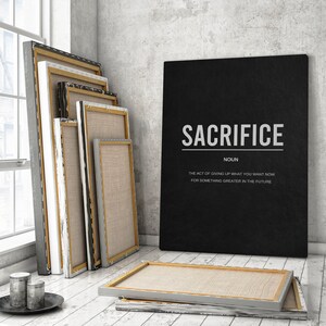 May include: A black canvas print with the word "SACRIFICE" in white text and the definition "The act of giving up what you want now for something greater in the future." The print is leaning against a stack of blank canvases.