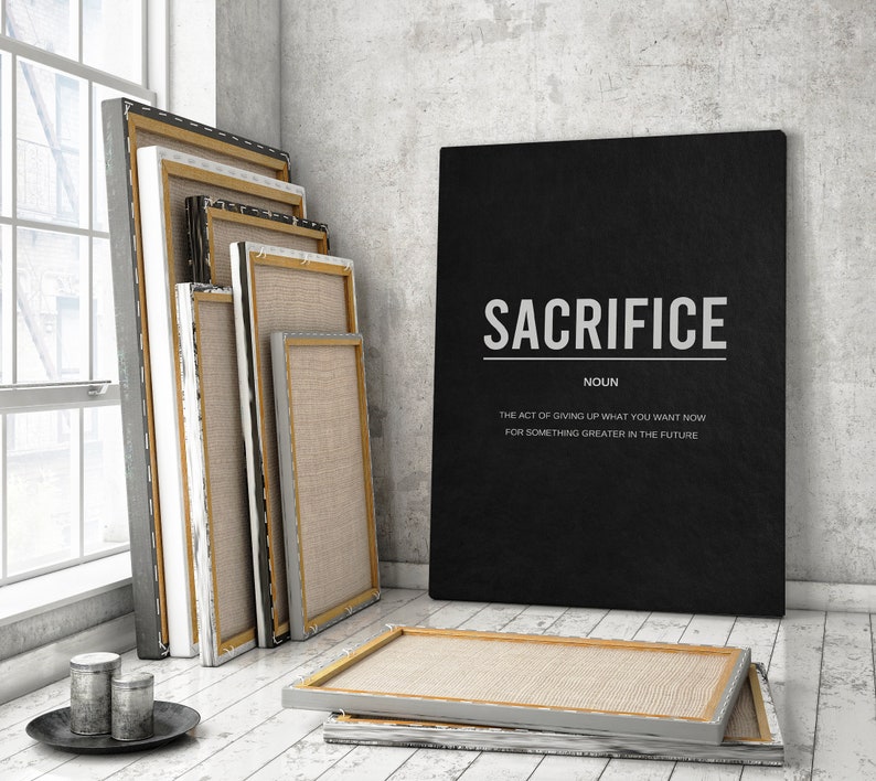 May include: A black canvas print with the word "SACRIFICE" in white text and the definition "The act of giving up what you want now for something greater in the future." The print is leaning against a stack of blank canvases.