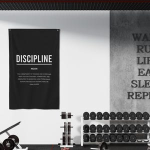 Pu&ograve; includere: Un poster motivazionale in bianco e nero con la parola "DISCIPLINE" in grandi lettere e una definizione della parola sotto. Il poster &egrave; appeso a un muro bianco in un ambiente di palestra. Il muro dietro il poster ha le parole "WAKE. RUN. LIFT. EAT. SLEEP. REPEAT." scritte in nero.
