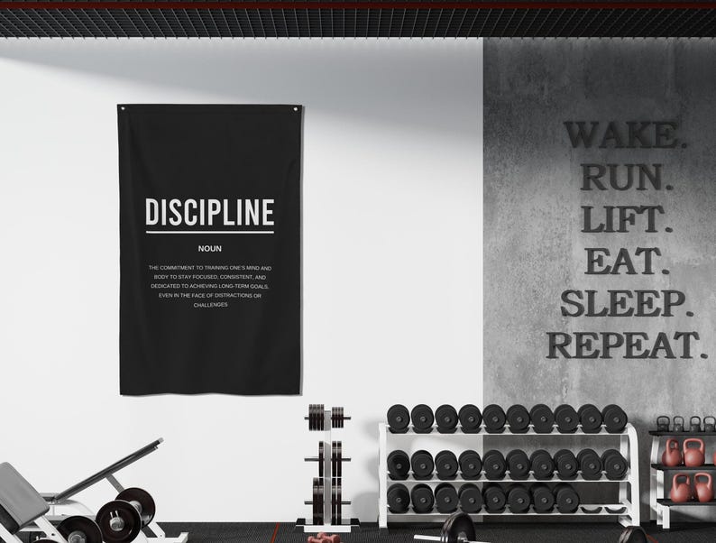 Pu&ograve; includere: Un poster motivazionale in bianco e nero con la parola "DISCIPLINE" in grandi lettere e una definizione della parola sotto. Il poster &egrave; appeso a un muro bianco in un ambiente di palestra. Il muro dietro il poster ha le parole "WAKE. RUN. LIFT. EAT. SLEEP. REPEAT." scritte in nero.