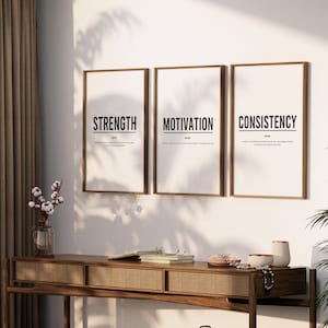 May include: Three framed prints with the words "Strength", "Motivation", and "Consistency" in black text on a white background. The prints are hanging on a white wall above a wooden console table with a woven rattan front.