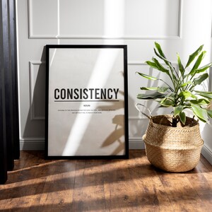 May include: A black framed poster with the word "CONSISTENCY" in bold white letters. The word "NOUN" is below the word "CONSISTENCY" in smaller white letters. Below the word "NOUN" is a definition of the word "CONSISTENCY" in smaller white letters. The poster is on a wooden floor with a green plant in a woven basket to the right.