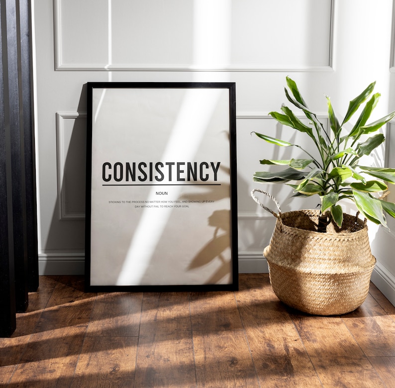 May include: A black framed poster with the word "CONSISTENCY" in bold white letters. The word "NOUN" is below the word "CONSISTENCY" in smaller white letters. Below the word "NOUN" is a definition of the word "CONSISTENCY" in smaller white letters. The poster is on a wooden floor with a green plant in a woven basket to the right.