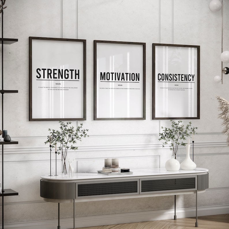 May include: Three black framed prints with white backgrounds and black text. The prints are hung on a white wall and read "Strength", "Motivation", and "Consistency".