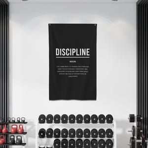 Pu&ograve; includere: Un poster motivazionale in bianco e nero con la parola "DISCIPLINE" in grandi lettere bianche. Il poster definisce la disciplina come "l'impegno di allenare la mente e il corpo per rimanere concentrati, coerenti e dedicati al raggiungimento di obiettivi a lungo termine, anche di fronte alle distrazioni della vita quotidiana."