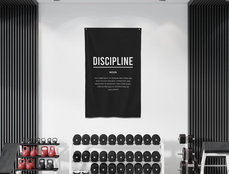 Pu&ograve; includere: Un poster motivazionale in bianco e nero con la parola "DISCIPLINE" in grandi lettere bianche. Il poster definisce la disciplina come "l'impegno di allenare la mente e il corpo per rimanere concentrati, coerenti e dedicati al raggiungimento di obiettivi a lungo termine, anche di fronte alle distrazioni della vita quotidiana."