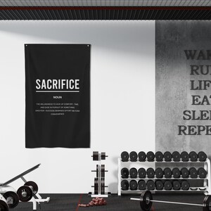 Pu&ograve; includere: Un poster motivazionale in bianco e nero con la parola "SACRIFICE" e la sua definizione. Il poster &egrave; appeso a un muro bianco in un ambiente di palestra. In primo piano ci sono pesi e una sbarra.