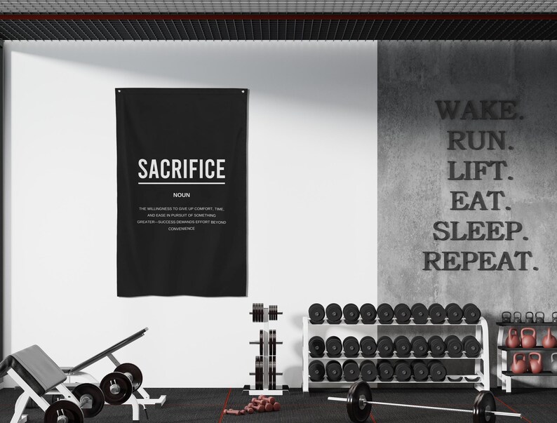 Pu&ograve; includere: Un poster motivazionale in bianco e nero con la parola "SACRIFICE" e la sua definizione. Il poster &egrave; appeso a un muro bianco in un ambiente di palestra. In primo piano ci sono pesi e una sbarra.
