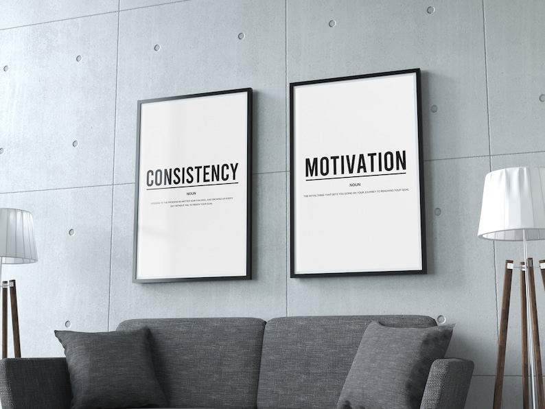 May include: Two black framed posters with white backgrounds and black text. The left poster says "CONSISTENCY" and the right poster says "MOTIVATION". The posters are hanging on a gray concrete wall above a gray couch.