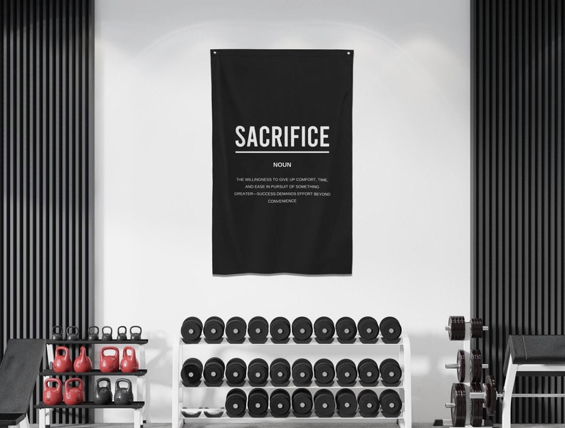 Pu&ograve; includere: Striscione motivazionale da parete per palestra in bianco e nero con la parola "SACRIFICE" in grandi lettere bianche. La definizione di sacrificio &egrave; scritta in lettere bianche pi&ugrave; piccole sotto la parola. Lo striscione &egrave; appeso a una parete bianca in un ambiente da palestra con pesi in primo piano.