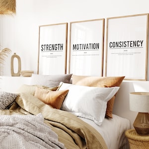 May include: Three framed prints with the words "Strength", "Motivation", and "Consistency" in black text on a white background. The prints are hanging on a white wall above a bed with brown bedding and pillows.