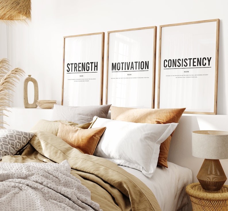 May include: Three framed prints with the words "Strength", "Motivation", and "Consistency" in black text on a white background. The prints are hanging on a white wall above a bed with brown bedding and pillows.