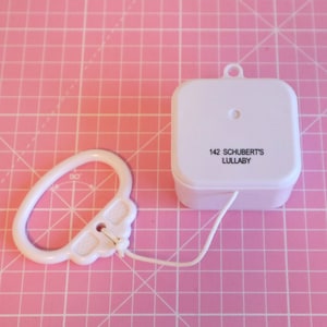 Może przedstawiać: Biała, nakręcana skrzynka muzyczna z sznurkiem do ciągnięcia. Skrzynka ma kwadratowy kształt i napis "142 SCHUBERT'S LULLABY" nadrukowany na górze.