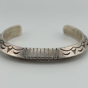 May include: A silver cuff bracelet featuring a central rectangular section with vertical grooves, flanked by engraved fan-shaped patterns on both sides. The metal has a polished finish with darkened recessed lines highlighting the intricate details.